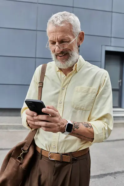 A bearded man smiles as he checks his smartphone while walking outside a modern building. — Stock Photo