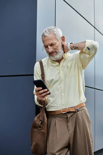 A bearded gay man with glasses stands outside near a modern building, checking his phone. — Stock Photo