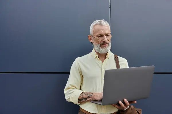 A mature man with a beard uses a laptop outside a modern building. — Stock Photo