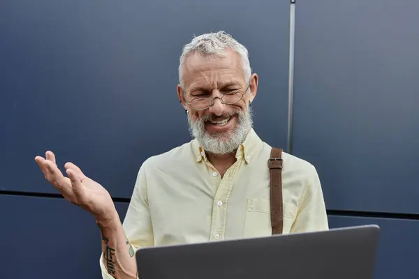 A bearded man in a yellow shirt and glasses smiles while using a laptop, with his left hand raised in the air. — Stock Photo