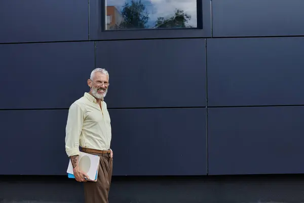 A mature gay man with a beard smiles as he stands outdoors near a modern building, holding books. — Stock Photo