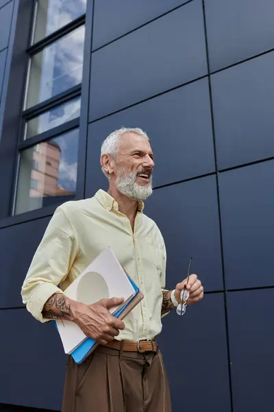 Un uomo di mezza età con la barba sorride mentre tiene libri e bicchieri. — Foto stock