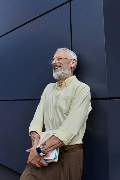 A middle-aged man with a beard laughs while leaning against a building, holding a stack of papers. — Stock Photo