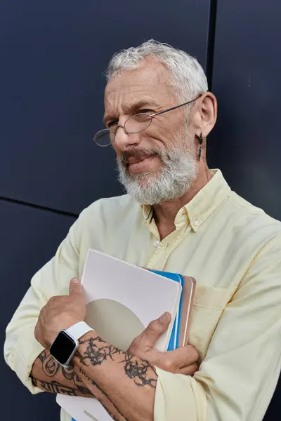 A mature gay man with a beard stands outside a modern building, holding books close to his chest. — Stock Photo