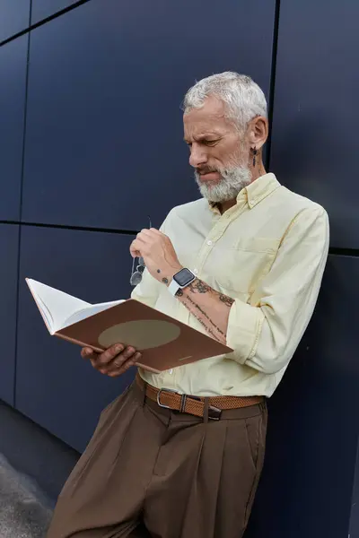 A bearded, middle-aged man in a yellow shirt reads a book while standing against a modern building. — Stock Photo