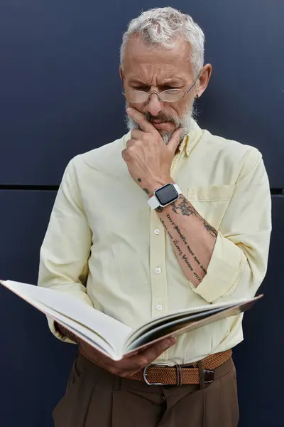 A mature man with a beard and glasses thoughtfully reads a book while standing near a modern building. — Stock Photo