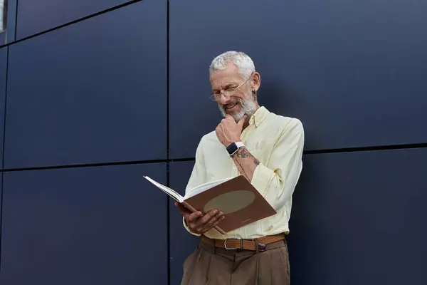 Зрілий гей-чоловік з бородою читає книгу, стоячи поруч з сучасною будівлею, задумливо торкаючись його підборіддя. — стокове фото