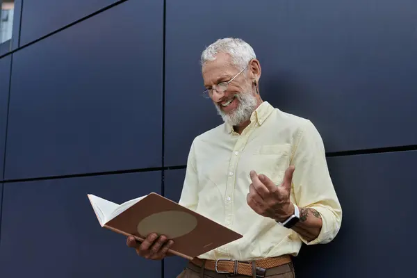 Ein bärtiger Mann im gelben Hemd lächelt, als er vor einem modernen Gebäude ein Buch liest. — Stockfoto