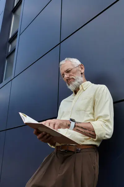 A middle-aged gay man with a beard reads a book while standing in front of a modern building. — Stock Photo