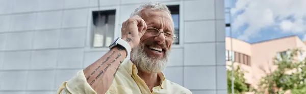 A mature man with a beard and glasses smiles broadly outdoors near a modern building. — Stock Photo