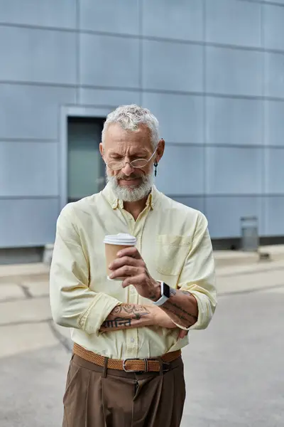 A mature gay man with a beard and glasses enjoys a cup of coffee outside a modern building. — Stock Photo