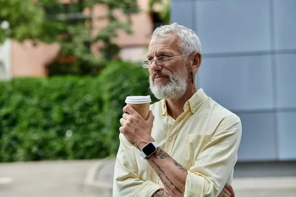 Middle-aged man with beard, glasses holds coffee, gazes at modern building, enjoying view — Stock Photo