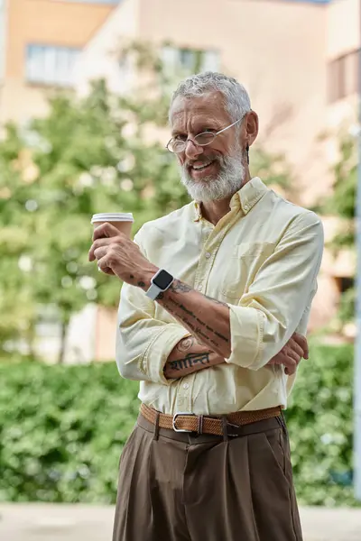 Un uomo maturo con la barba sta fuori, sorridendo mentre tiene in mano una tazza di caffè. — Foto stock