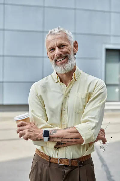 Un homme mûr avec une barbe et un sourire chaleureux se tient dehors, tenant une tasse de café. Il porte une chemise jaune clair et un pantalon marron. — Photo de stock