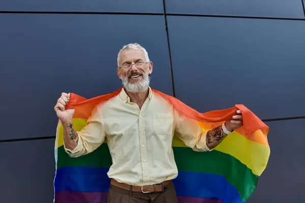 A mature man with a beard and glasses smiles while holding a rainbow pride flag. — Stock Photo