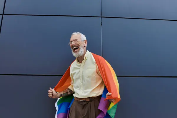 Un uomo gay maturo con la barba ride mentre tiene una bandiera arcobaleno fuori da un edificio moderno. — Foto stock