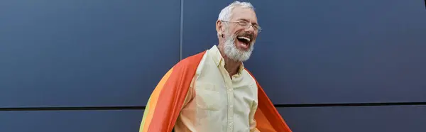 A mature man with a beard laughs outside a modern building, wearing a pride flag like a cape. — Stock Photo