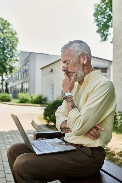 A mature man with a beard sits on a bench outside a modern building, using his laptop and deep in thought. — Stock Photo