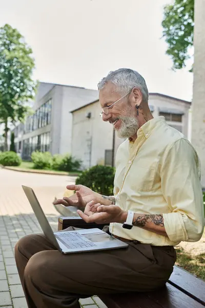 A mature man with a beard sits on a bench, smiling as he uses his laptop. He is wearing a light yellow shirt and has a tattoo on his arm. — Stock Photo