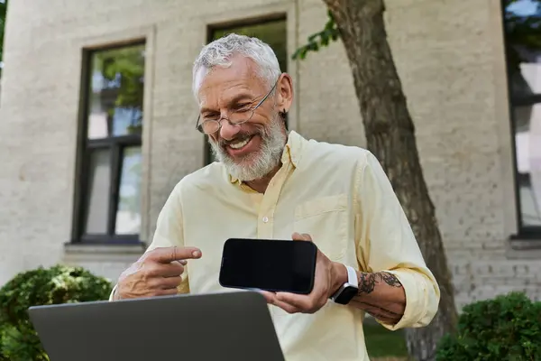 Un homme gay mature avec une barbe sourit et pointe vers un écran de téléphone tout en étant assis à l'extérieur avec un ordinateur portable. — Photo de stock