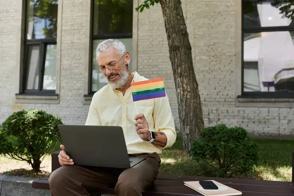 A mature gay man with a beard sits outdoors near a modern building, holding a rainbow flag and using a laptop. — Stock Photo