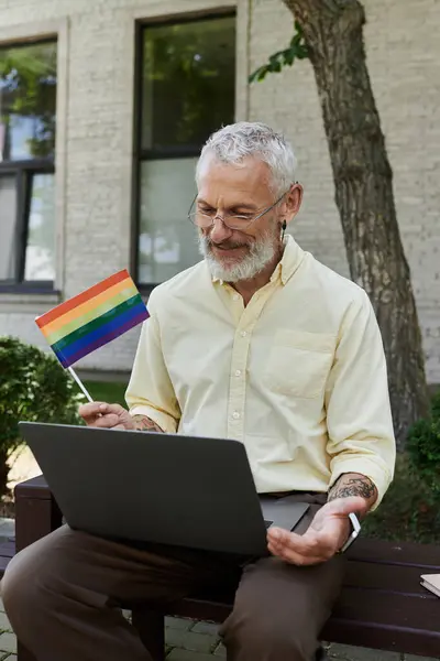 Ein schwuler Mann mittleren Alters mit Bart und Brille sitzt auf einer Bank vor einem modernen Gebäude und lächelt, während er einen Laptop benutzt.. — Stockfoto