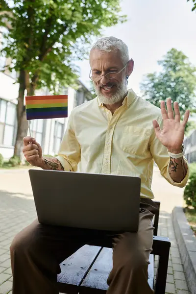 Um homem gay de meia-idade com uma barba sorri e acena enquanto ele chats de vídeo em seu laptop, segurando uma bandeira do arco-íris. — Fotografia de Stock