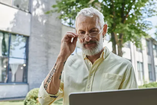 A mature man with a beard and glasses adjusts his spectacles while looking at a laptop outside a modern building. — Stock Photo