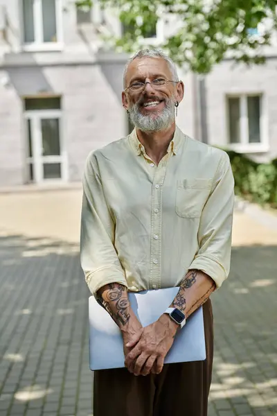 A mature, bearded man with glasses smiles while holding a laptop outdoors. — Stock Photo