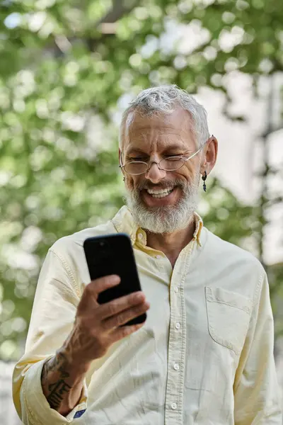 A mature man with a beard and glasses smiles at his phone outdoors. — Stock Photo