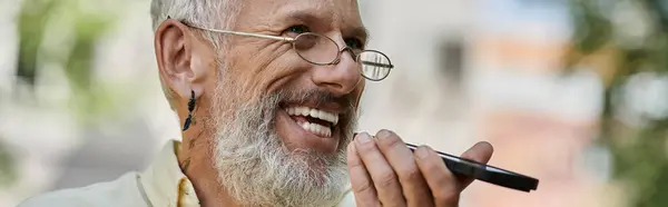 A mature man with a beard and glasses smiles while using his phone outside a modern building. — Stock Photo