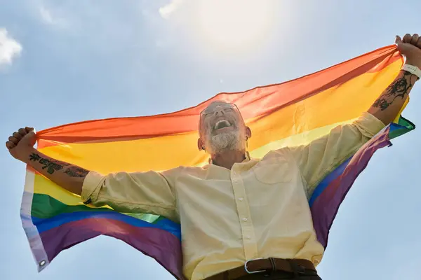 A man with a beard and tattoos holds a rainbow pride flag high in the air, celebrating with a joyful smile. — Stock Photo