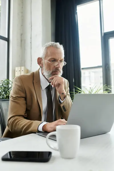 A middle-aged lgbtq man with a beard, wearing glasses and a suit jacket, sits at a desk in front of a laptop and contemplates — Stock Photo