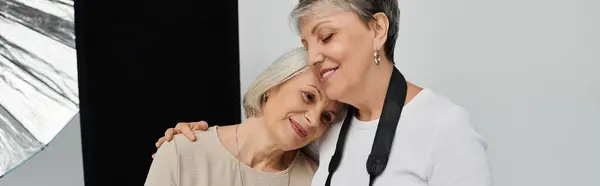 A lesbian couple poses in a studio, one woman wearing a camera strap, gently embraces her partner. — Stock Photo