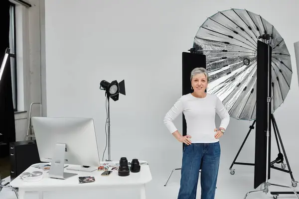 A mature woman, dressed in a white shirt and blue jeans, stands confidently in a photo studio. — Stock Photo