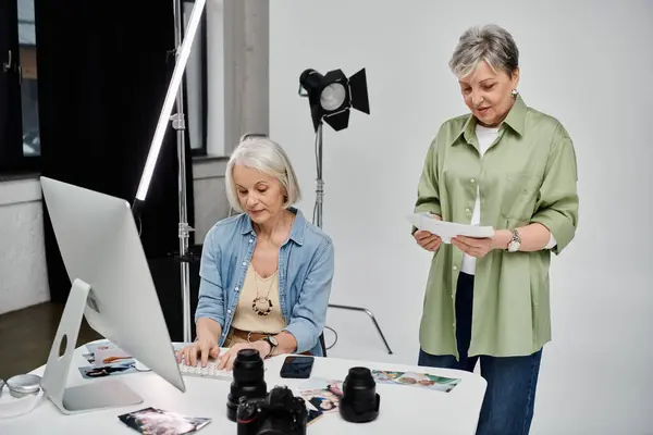A lesbian couple is in a photo studio. One woman is reviewing photos on a computer while the other woman holds up photographs for review. — Stock Photo