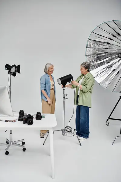 Dos mujeres lesbianas maduras, una posando como modelo, la otra como fotógrafa, durante una sesión de fotos en un estudio. - foto de stock