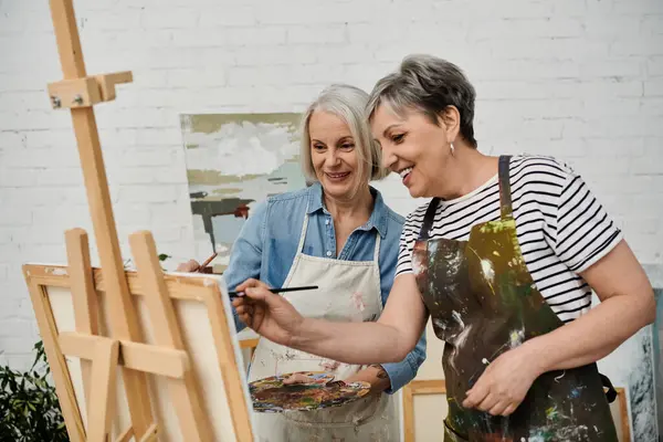 Two women in aprons paint together in a studio. One paints with a brush, focused; the other smiles behind her — Stock Photo