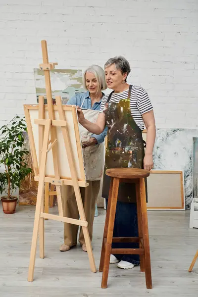 Two women, wearing paint-splattered aprons, work together on a canvas in a bright art studio. — Stock Photo