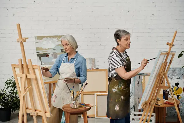 Two women, wearing paint-splattered aprons, stand at easels in a white brick studio, focused on their art. — Stock Photo