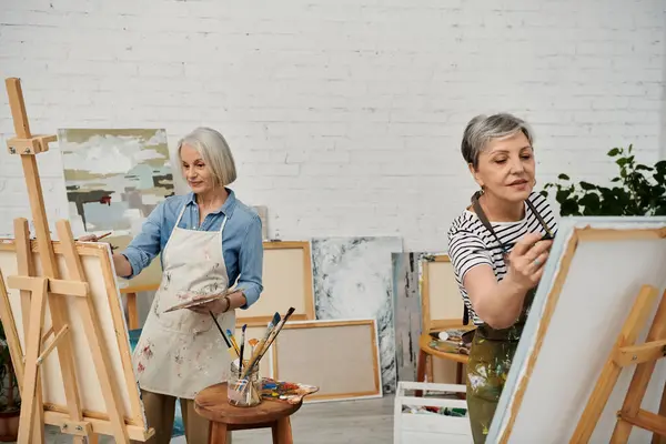 Two mature women, dressed in casual attire, paint at easels in an art studio filled with canvases and supplies. — Stock Photo