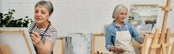 Two mature women paint side-by-side in an art studio, focusing on their craft with paintbrushes and canvases. — Stock Photo