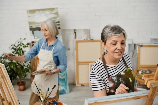 Two older women paint canvases in a well-lit art studio, surrounded by easels and art supplies. — Stock Photo