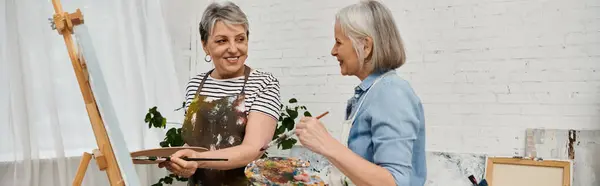 Zwei reife Frauen stehen in einem Kunstatelier, eine hält Pinsel und Palette in der Hand, die andere lächelt und hält einen Pinsel in der Hand.. — Stockfoto