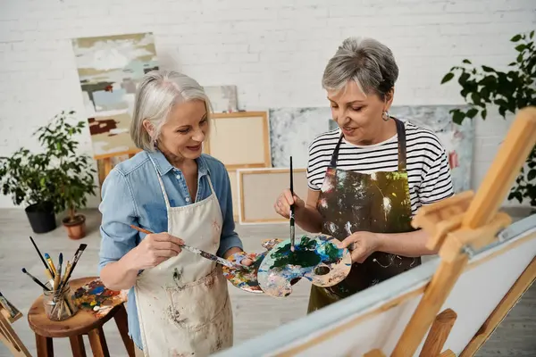 Two women stand together in an art studio, holding paintbrushes and a palette, engaged in a creative and vibrant painting session. — Stock Photo