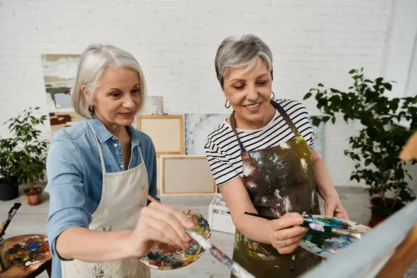Two mature women, dressed in aprons, are painting with brushes and palettes in an art studio. — Stock Photo