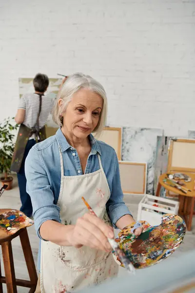 An older woman paints with focus and passion in her art studio, surrounded by other canvases and art supplies. — Stock Photo