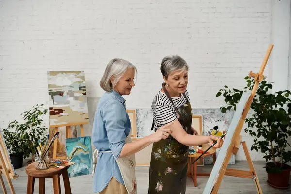 Two mature women in an art studio, one painting and the other giving guidance. — Stock Photo