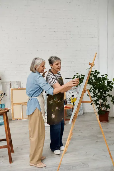 Two mature women, in an art studio, stand in front of an easel, with one woman painting. — Stock Photo