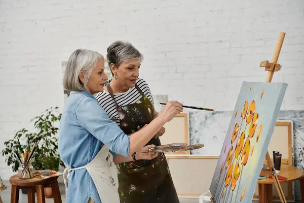 Deux femmes peignent une toile ensemble dans un atelier d'art, une femme tenant un pinceau et l'autre regardant l'œuvre. — Photo de stock